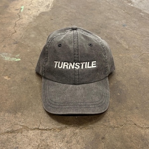 May include: A charcoal gray baseball cap with the word "TURNSTILE" embroidered in white across the front. The cap has a curved brim and a six-panel design, with a slightly faded, vintage look.