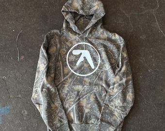 May include: A camouflage-patterned hoodie with a white circular logo on the front. The hoodie features a drawstring hood and a kangaroo pocket. The camouflage design incorporates shades of gray, brown, and green.