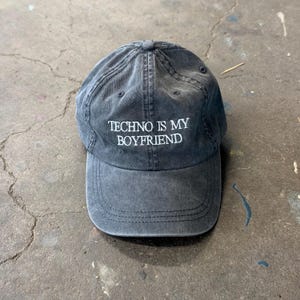 以下が含まれることがあります： フロントパネルの中央に白い刺繍で「TECHNO IS MY BOYFRIEND」と書かれた、ウォッシュ加工のチャコールグレーのベースボールキャップ。キャップにはカーブしたつば、上部のボタン、通気用のステッチ入りアイレットが付いています。