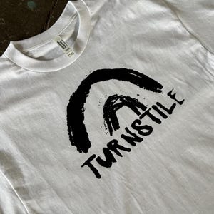 May include: White t-shirt with a black graphic design. The design features a stylized arch and the word "TURNSTILE" in a bold, handwritten font. The shirt is made of a soft material and has a crew neck.