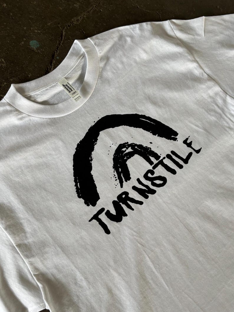 May include: White t-shirt with a black graphic design. The design features a stylized arch and the word "TURNSTILE" in a bold, handwritten font. The shirt is made of a soft material and has a crew neck.