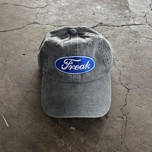 以下が含まれることがあります： カーブしたつばが付いたグレーの野球帽。「Freak」の文字が白い文字で青い楕円の中に刺繍されています。帽子は柔らかく、使い古された素材で作られています。
