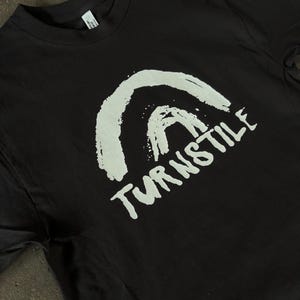May include: Black t-shirt featuring a white, brushstroke-style graphic of an arch and the word "TURNSTILE" in a handwritten font. The shirt has a crew neck and short sleeves. The fabric appears to be a soft cotton blend.