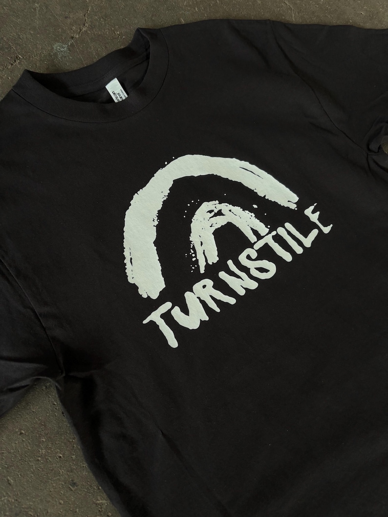 May include: Black t-shirt featuring a white, brushstroke-style graphic of an arch and the word "TURNSTILE" in a handwritten font. The shirt has a crew neck and short sleeves. The fabric appears to be a soft cotton blend.