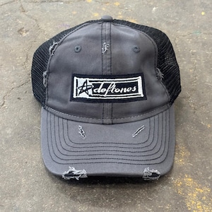 May include: Gray trucker hat with a black mesh back and a distressed brim. The hat features a rectangular patch with the band name "Deftones" in white lettering.