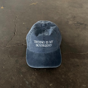 Puede incluir: Gorra de béisbol azul marino con el texto blanco "TECHNO IS MY BOYFRIEND". La gorra tiene una visera curva y un cierre de correa de tela. El sombrero está hecho de un material suave y desgastado.