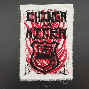 May include: A white fabric patch with a red and black design. The patch features the words "CHINGA" and "MIERA" in bold, stylized lettering above a black shield emblem with a star. The background has red swirls.