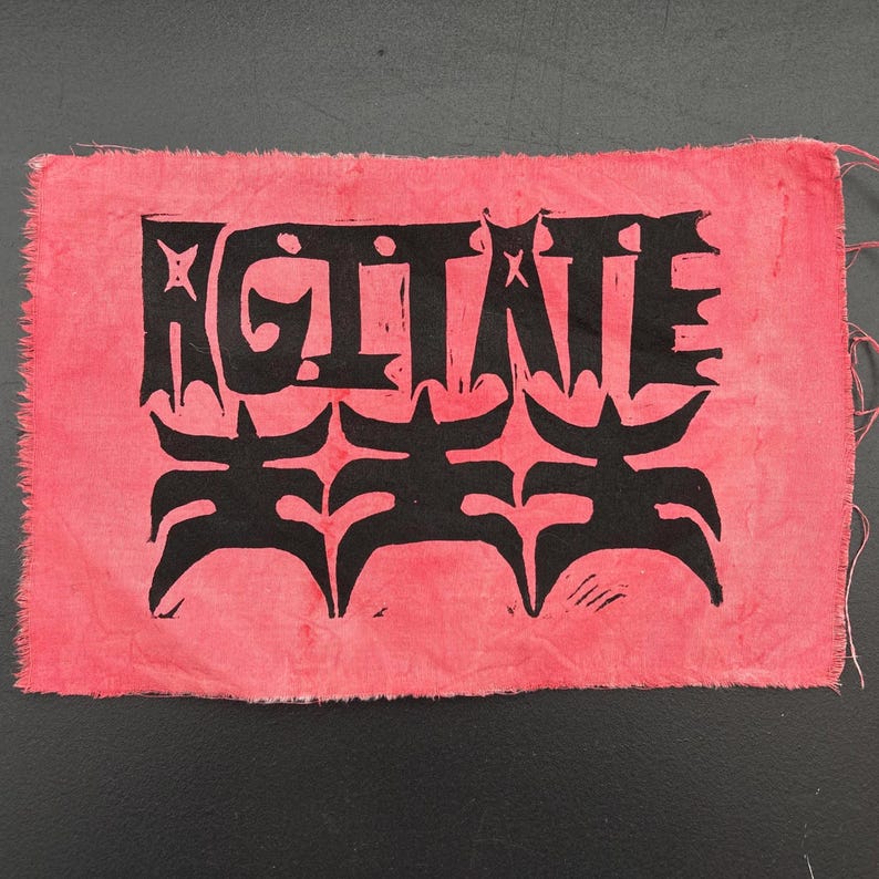 May include: A pink fabric with the word "AGITATE" in black block letters. Below the word are three stylized black figures. The fabric has a rough, textured appearance with frayed edges.