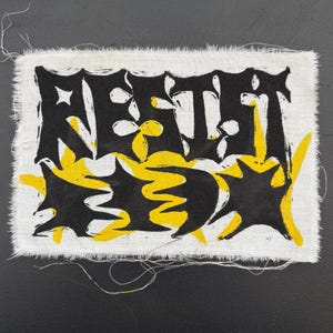 Op de afbeelding: Een rechthoekige lap witte stof met gerafelde randen. De lap toont het woord "REST" in zwarte, gestileerde letters, met gele accenten en vormen. De achtergrond is donkergrijs.