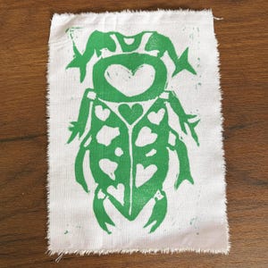 May include: A green insect print on a white fabric square. The insect design features a heart-shaped head and heart-shaped details on its body. The fabric has a raw, unfinished edge.