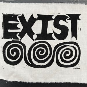 Könnte beinhalten: Ein weißes Stoffquadrat mit dem Wort "EXIST" in fetten, schwarzen, stilisierten Buchstaben. Darunter befinden sich drei schwarze Spiralmotive. Die Kanten des Stoffes sind ausgefranst.
