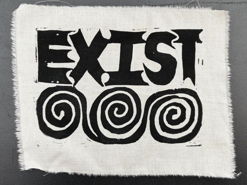 May include: A white fabric square with the word "EXIST" in bold, black, stylized letters. Below the word are three black spiral designs. The edges of the fabric are frayed.