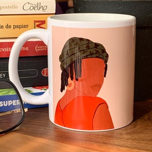 May include: White ceramic mug featuring an illustration of a person with a headscarf and orange top. The design is set against a light pink background. The mug has a white handle and is sitting on a wooden surface.