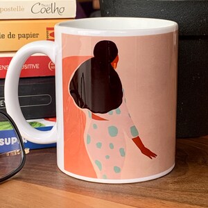 May include: White ceramic mug featuring an illustration of a person in a polka-dot outfit with a dark hairstyle. The background is a blend of peach and orange hues. The mug has a white handle and is placed on a wooden surface.