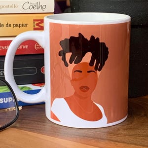 May include: White ceramic mug featuring a stylized illustration of a woman with dark hair and a white top against a terracotta background. The mug has a curved handle and is likely used for beverages.