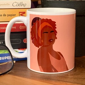 May include: White ceramic mug featuring a stylized illustration of a woman with dark skin, wearing an orange headband and a peach-colored top. The background is a soft peach color. The mug has a white handle.