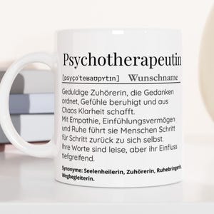 Könnte beinhalten: Weiße Keramik-Tasse mit dem Wort "Psychotherapeutin" in Schwarz aufgedruckt. Die Tasse enthält auch eine Definition auf Deutsch. Die Tasse steht auf einer weißen Oberfläche mit einem Stapel Bücher und einer dekorativen Vase im Hintergrund.