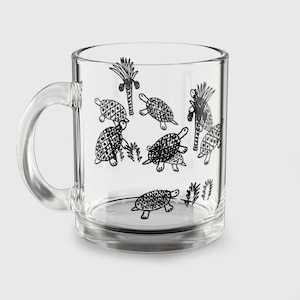 May include: Clear glass mug with a handle, adorned with black line drawings of turtles and palm trees. The turtles feature patterned shells and are set amongst stylized plants. The mug is cylindrical with a clear handle.