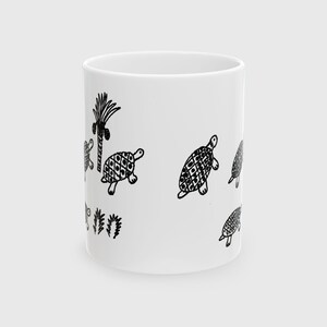 May include: White ceramic mug with a black illustration of turtles and a palm tree. The turtles have patterned shells. The mug is cylindrical and has a simple, clean design.