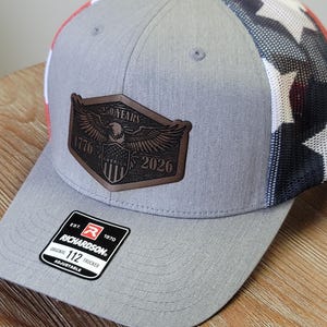 May include: A grey trucker hat featuring a brown leather patch with an eagle, a shield, and the text 250 YEARS 1776-2026 AMERICA. The hat has a mesh back with a red, white, and blue star pattern and a black sticker on the brim reading RICHARDSON 112.
