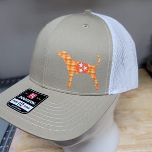 May include: A tan and white trucker hat with a dog silhouette design. The dog is orange and yellow plaid with three white stars. The hat has a curved brim and a white mesh back. The hat has the text "RICHARDSON" and "112 TRUCKER" on the front.