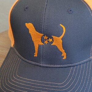 May include: A navy blue baseball cap with an orange mesh back and brim. The cap features an embroidered orange silhouette of a dog with stars. The stitching on the brim is orange.