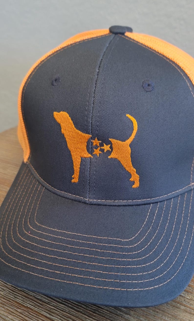 May include: A navy blue baseball cap with an orange mesh back and brim. The cap features an embroidered orange silhouette of a dog with stars. The stitching on the brim is orange.