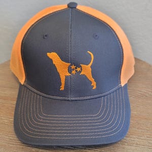 May include: A navy blue and orange trucker hat featuring an embroidered orange dog silhouette with a Tennessee state flag design. The hat has a mesh back and a curved brim with orange stitching.
