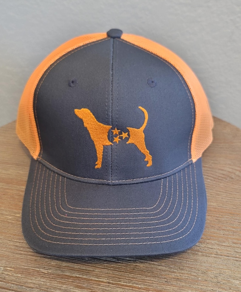 May include: A navy blue and orange trucker hat featuring an embroidered orange dog silhouette with a Tennessee state flag design. The hat has a mesh back and a curved brim with orange stitching.