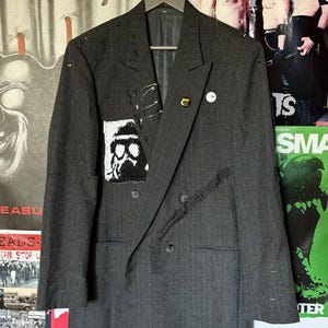 May include: A charcoal gray pinstriped double-breasted blazer featuring a white square patch with a black graphic print, multiple silver safety pins, a small circular pin, and decorative black stitching across the front panel.