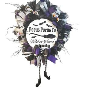 Könnte beinhalten: Ein Halloween-Kranz mit einem runden Schild mit der Aufschrift "Hocus Pocus Co" mit Fledermäusen und einer Besen-Grafik. Der Kranz ist mit schwarzen, weißen und lila Bändern und Hexenbeinen mit gestreiften Strümpfen verziert.