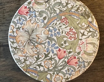 Puede incluir: Posavasos redondo de cerámica con un diseño floral. El diseño presenta flores y follaje estilizados en tonos rosa, azul, verde y naranja sobre un fondo blanco. El posavasos tiene aproximadamente 10 cm de diámetro.