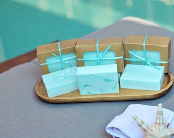 May include: A wooden tray holds three bars of light blue soap and three small brown boxes tied with light blue ribbon. The soap has a marbled appearance. A white towel and a starfish are in the foreground.
