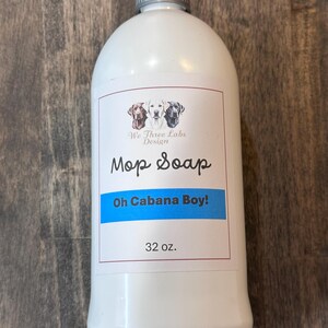 Könnte beinhalten: Weiße Plastikflasche "Mop Soap" mit silbernem Verschluss. Das Etikett zeigt den Text "We Three Labs Design", "Mop Soap" und "Oh Cabana Boy!" in Blau. Die Flasche enthält 946 ml Produkt.