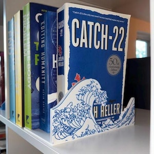Puede incluir: Una estantería con varios libros, incluyendo "Catch-22" de Joseph Heller. El libro está sostenido por un sujetalibros blanco con un diseño de ola azul. Otros libros incluyen títulos como "Editing Humanity" y "The Three-Body Problem".