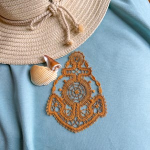 May include: A tan sun hat with a rope tie, a seashell, and a detailed orange lace anchor design on a light blue surface. The anchor design features a ship's wheel and intricate openwork.