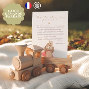 May include: A wooden toy train with a card that reads "Will you be my godfather?" The card features a bear illustration. The train is light brown and sits on a white blanket, with a blurred outdoor background.