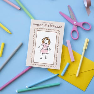 May include: A light beige card with the text "Portrait de ma Super Maîtresse" and a drawing of a person in a pink dress. The card is surrounded by pink scissors, pens, a yellow envelope, and tape on a blue surface.