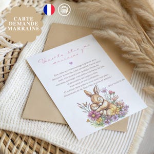 May include: A greeting card with the text "Veux-tu être ma marraine?" and a drawing of a sleeping bunny surrounded by flowers. The card is on a beige envelope, with the words "CARTE DEMANDE MARRAINE" on the top left.
