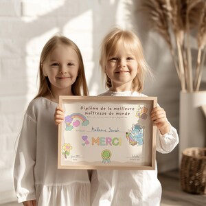 May include: Two young children holding a framed certificate. The certificate has a light wood frame and features colorful illustrations of a rainbow, flowers, and a cartoon hippo. Text on the certificate reads "Diplôme de la meilleure maîtresse du monde" and "Madame Sarah".