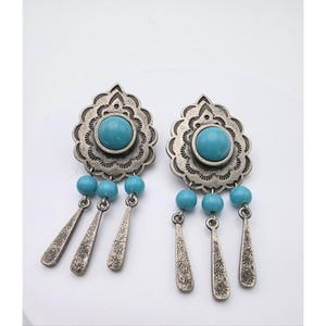 May include: A pair of silver-coloured earrings featuring a turquoise stone centre and turquoise bead accents. The earrings have a decorative, scalloped design around the central stone and dangling, teardrop-shaped charms.
