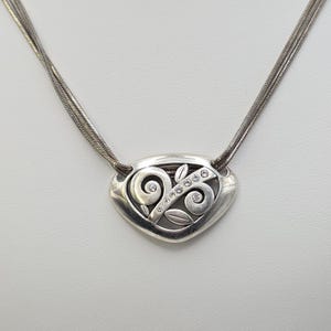 May include: Silver-toned necklace featuring a heart-shaped pendant with an openwork design. The pendant is adorned with small, clear crystals and swirling patterns. The necklace has a multi-strand chain.