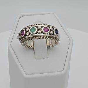 May include: A silver ring with a braided band and a row of alternating colored gemstones. The gemstones include red, green, and blue stones. The ring is displayed on a white surface.