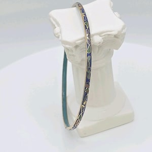 Pode incluir: Uma pulseira fina, dourada, com interior em esmalte azul. O exterior apresenta um padrão repetido de desenhos florais azuis, verdes e dourados. A pulseira é exibida em um pedestal branco.