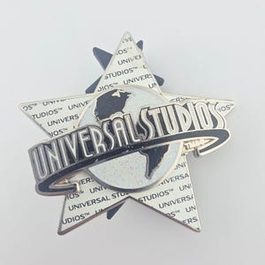 May include: A metal pin featuring a white star background patterned with the repeating text Universal Studios. Centered on the star is a globe graphic with a black landmass and a white glitter ocean, overlaid by a black Universal Studios logo.
