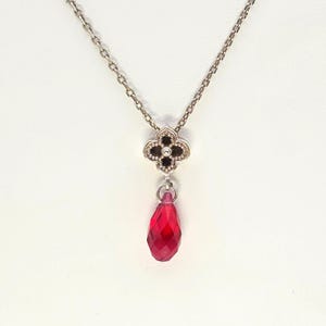 May include: Silver-toned necklace featuring a four-leaf clover pendant with black accents and a central gem. A faceted, teardrop-shaped, red gemstone hangs below the clover. The chain is delicate and the background is white.