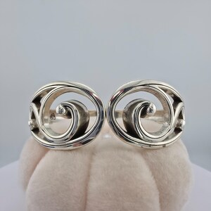 May include: A silver cuff bracelet featuring two symmetrical, open-work circular motifs on each end. Each circular element contains a central, inward-curving spiral design. The metal has a polished, reflective finish with smooth, rounded edges.