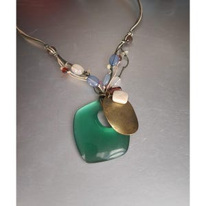 Puede incluir: Un collar plateado con un colgante verde cuadrado con un agujero. El collar presenta un colgante ovalado color latón y cuentas de colores en tonos azules, blancos y rojos. El collar es una pieza de joyería única.