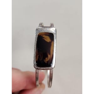Puede incluir: Una pulsera rígida de tono plateado con una pieza central rectangular que presenta una incrustación con patrón de carey en marrón oscuro y beige. La palabra GUESS está grabada en letras mayúsculas pequeñas en el marco plateado.
