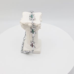 Könnte beinhalten: Ein silberfarbenes Armband mit Eidechsen-Anhängern mit farbigen Akzenten. Das Armband wird auf einem weißen Sockel präsentiert, der das filigrane Design und die leuchtenden Farben der Edelsteine hervorhebt.